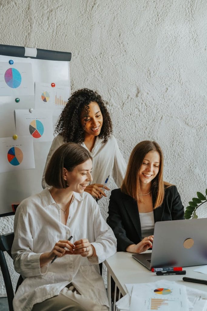 women-in-the-office-looking-at-laptop-screen-smiling-8145247 Three businesswomen collaborate with graphs and a laptop in a modern office setting.
